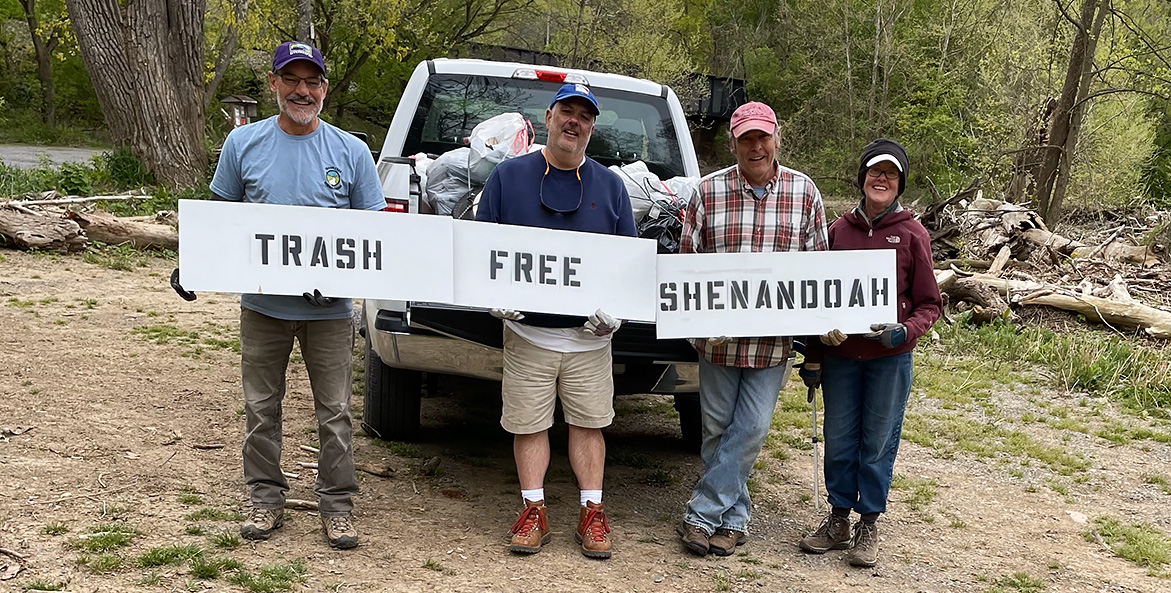 Four people stand behind a white truck filled with white trash bags, holding three signs that say Trash Free Shenandoah.