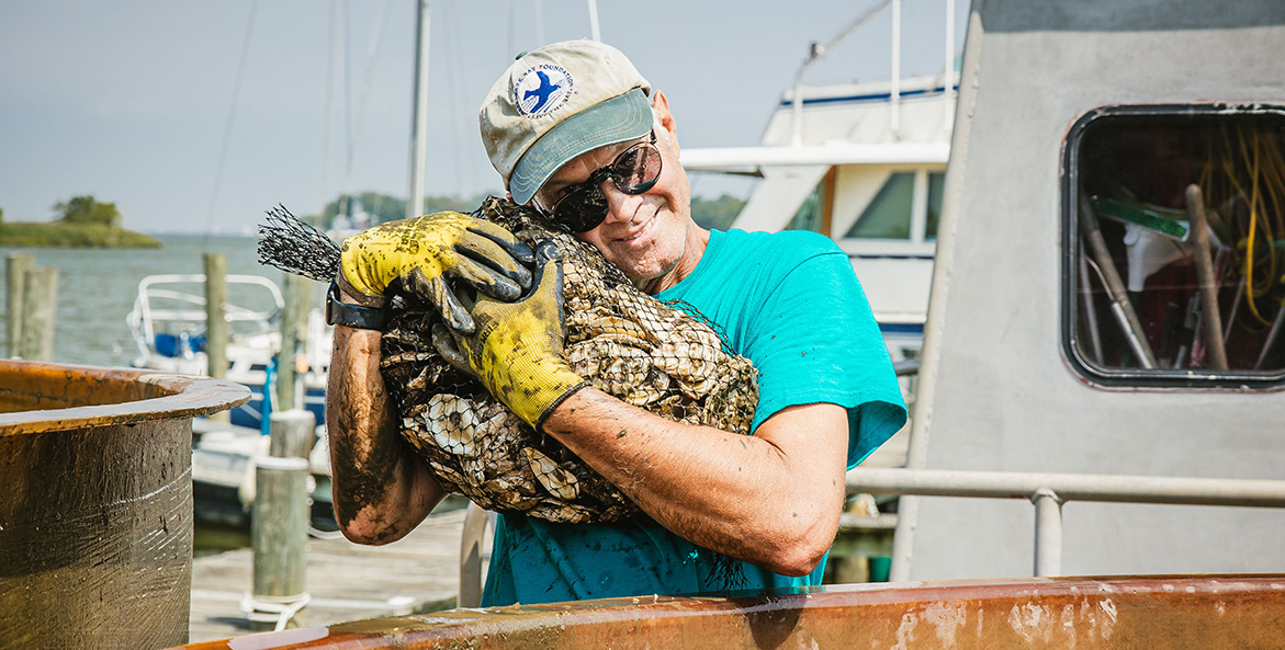 A man with a CBF cap and work gloves hugs a bag of oyster shell.