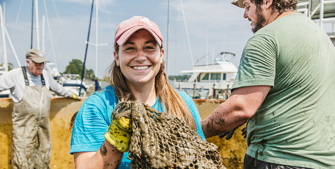 A smiling woman holds a mud-covered net bag of oysters.