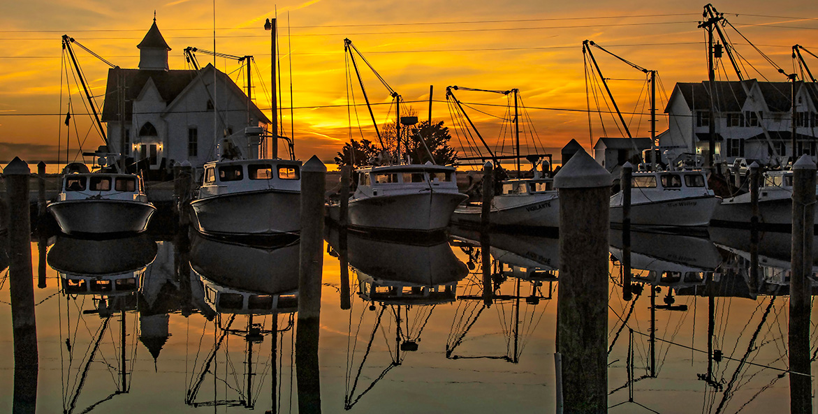 The sun sets, casting an orange glow across the sky and water, where six fishing boats are docked.