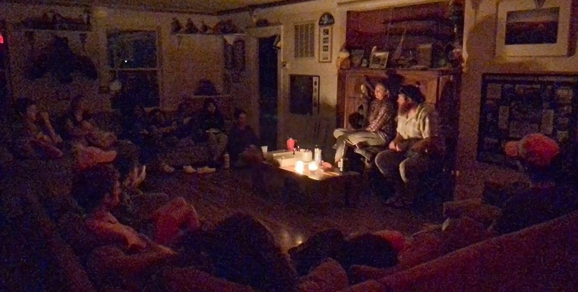 Two people sit in the light of an oil lamp and candle light, surrounded by others seated on couches in the shadows.