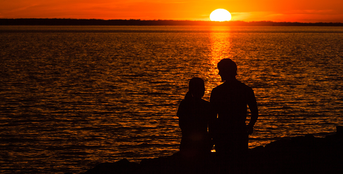 A woman and man are silhouetted against the orange light of the sun setting on the other side of the river.