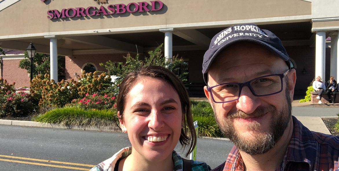 Two people stand in front of the entrance to a building with a sign reading Smorgasbord.