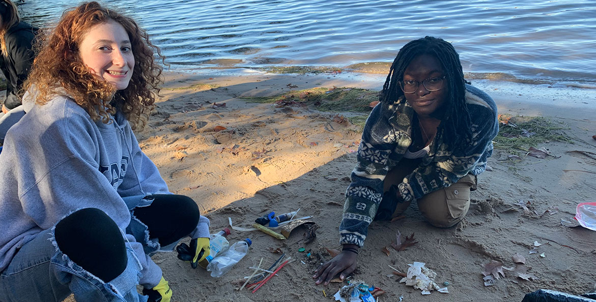 Two young girls kneel on a beach with a collection of trash in front of them