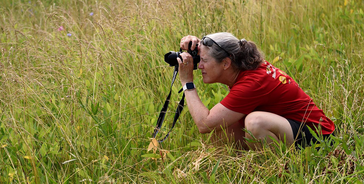 A woman crouches in the grass, looking through and focusing a 35mm camera.