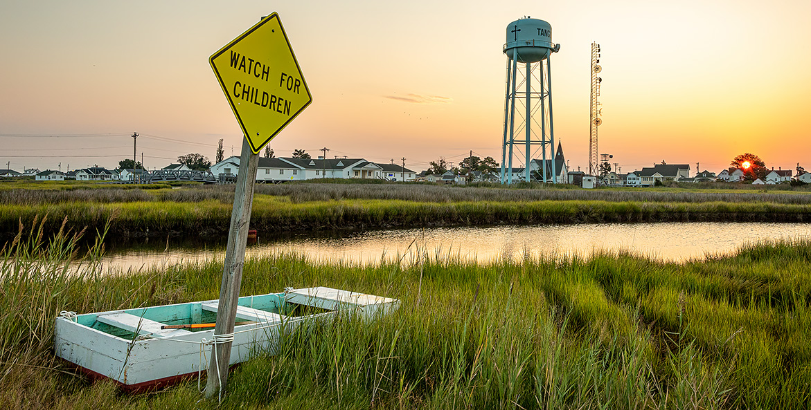 A skiff is tied up to a sign that reads Watch for Children on a marsh across an inlet from buildings and a water tower at sunrise.