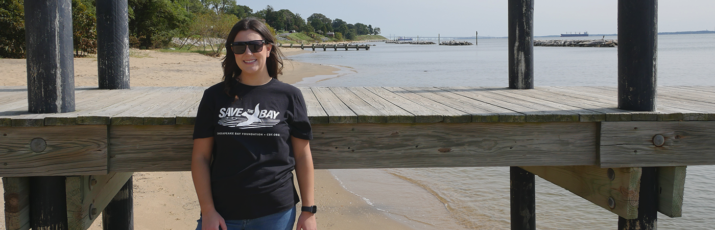 Woman wearing a black Save the Bay t-shirt stands by a pier on the beach.